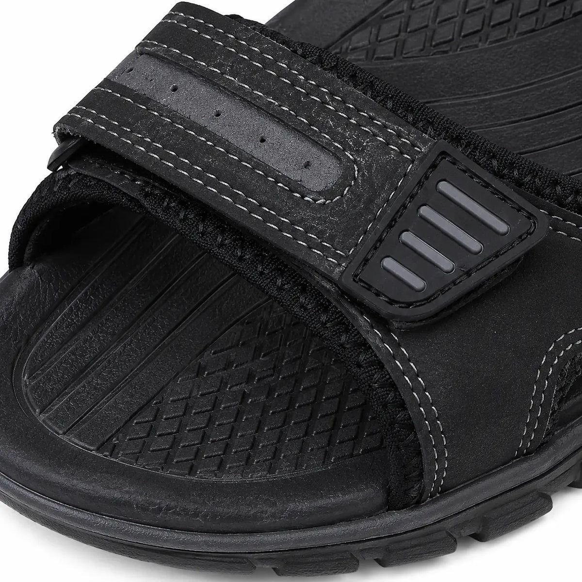 A close-up of a black slide sandal showing the textured sole, hook-and-loop strap, and contrast stitching details.