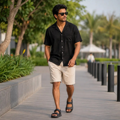 A man walking outdoors on a paved path wearing a black short-sleeve button-up shirt, beige shorts, black sandals, and sunglasses.